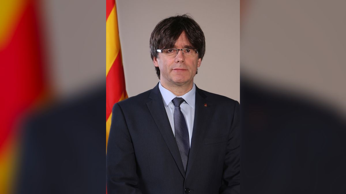 Retrato oficial de Carles Puigdemont con traje oscuro, gafas y bandera catalana al fondo