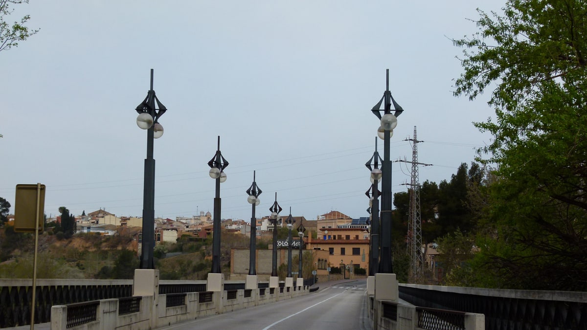 Puente con farolas de hierro negro sobre el río, con el casco urbano de Sant Sadurní al fondo.