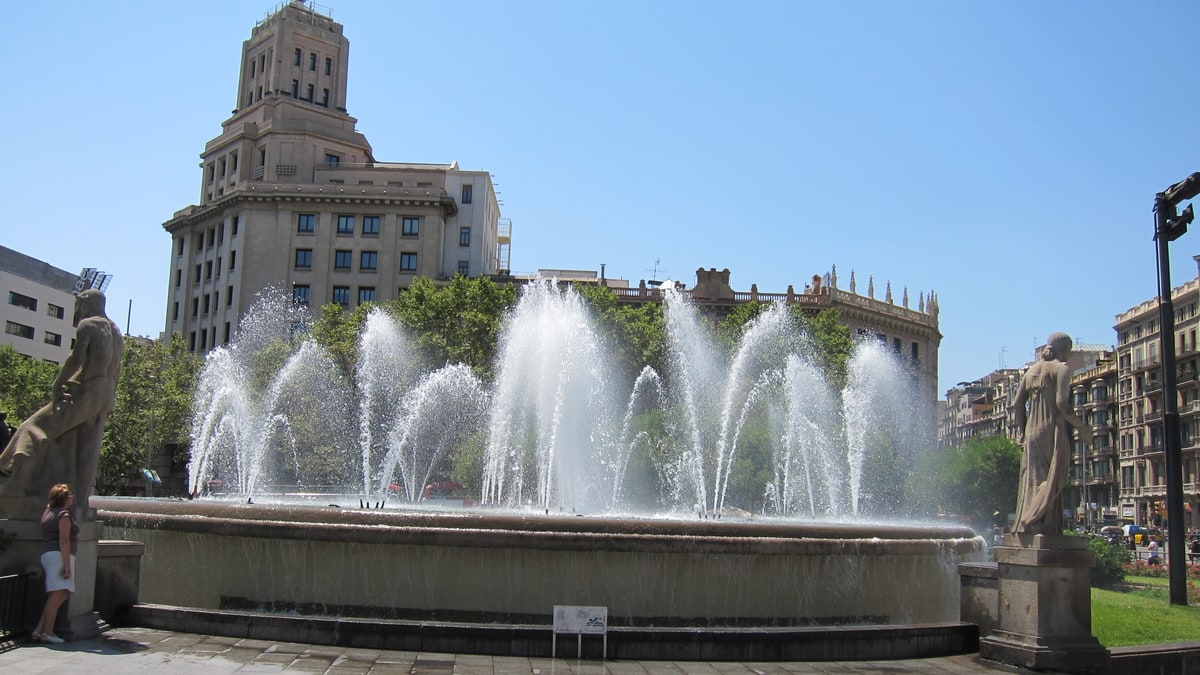 Fountain with water jets in Plaza de Catalunya, Barcelona, with historic buildings in the background.