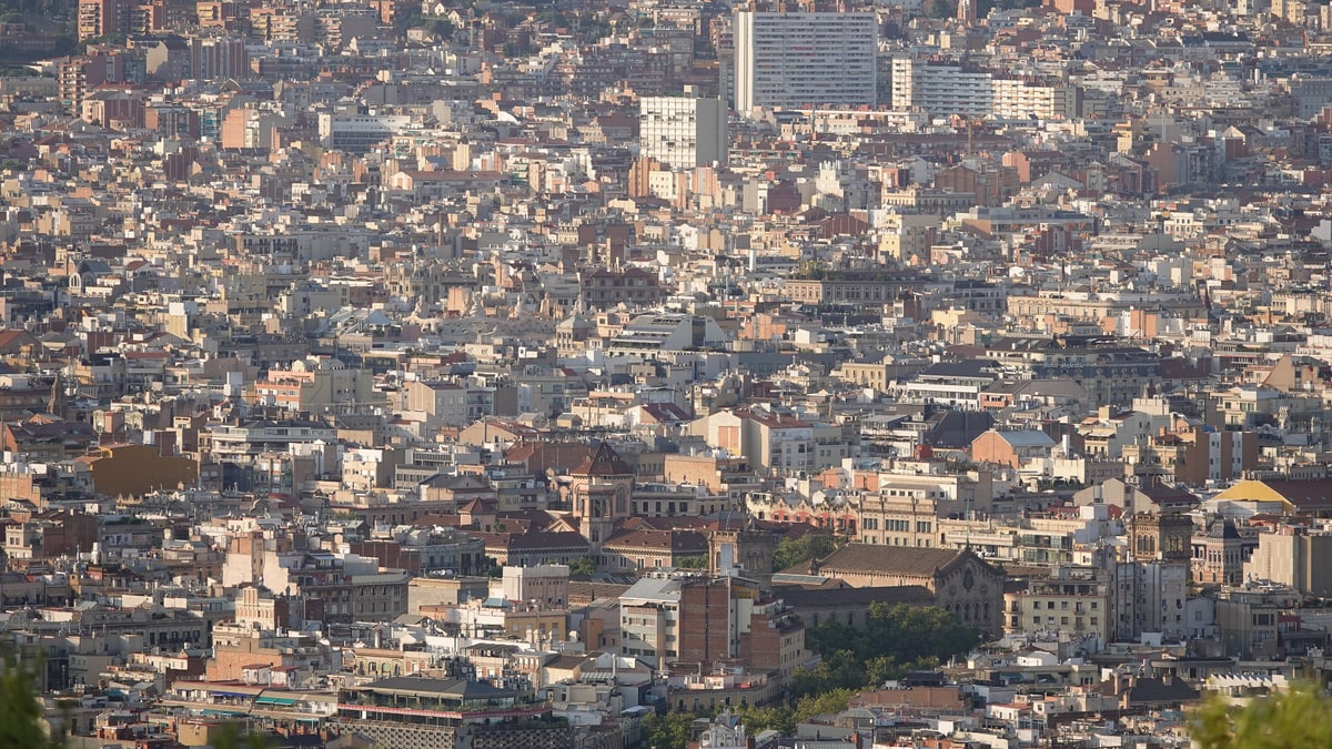 Vista aérea del denso entramado urbano de Barcelona con edificios de varias alturas y montañas al fondo.