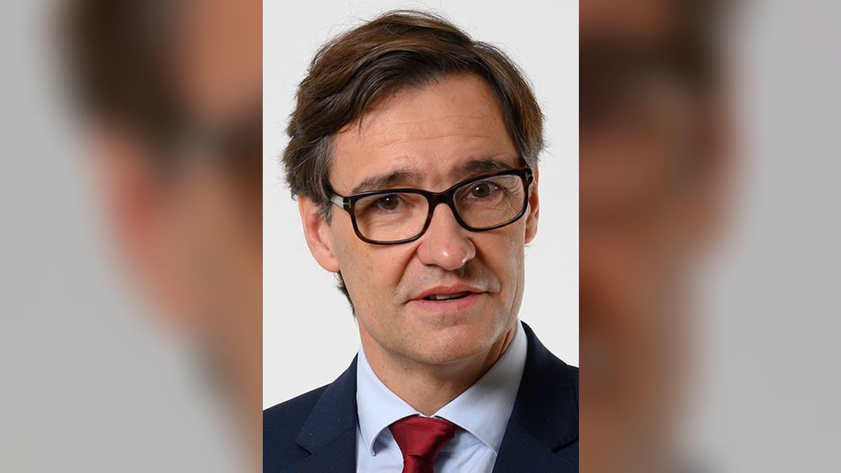 Retrato de Salvador Illa con gafas, traje oscuro y corbata roja, mirando a cámara sobre fondo claro
